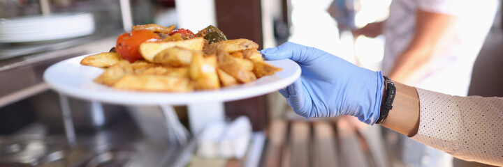 Female hand in blue medical glove hold white plate with fried potatoes and stewed vegetables. Self service restaurant with tray.