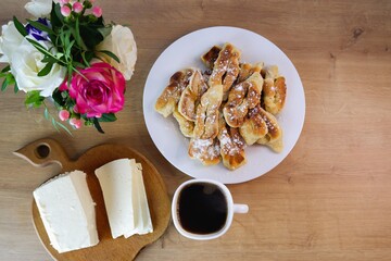 Home-baked brushwood during quarantine.  Simple food. Homemade feta cheese, a cup of coffee on a wooden table top. Concept on the background of a white brick wall and a bouquet of roses.