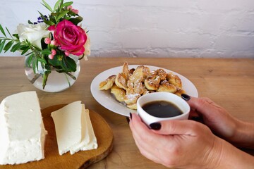 Cup of coffee in female hands, close-up. Homemade baked goods during quarantine.  Homemade feta cheese on a wooden tabletop. Concept on the background of a white brick wall and a bouquet of roses.