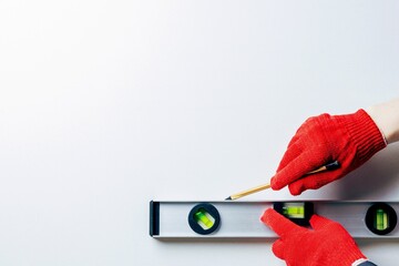 The hands of a worker in red protective gloves hold a metal spirit level and a pencil against a white wall. Home master concept