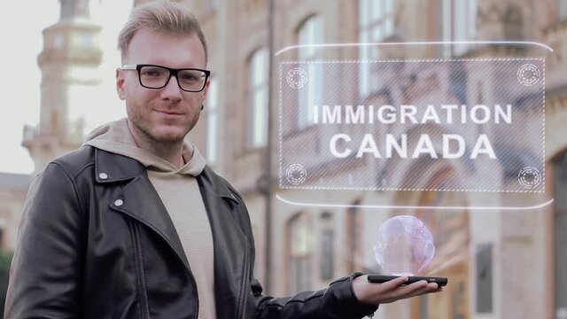 Smart young man with glasses shows a conceptual hologram Immigration Canada. Student in casual clothes with future technology mobile screen on university background