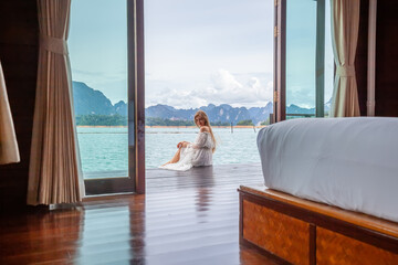Travel Woman in White Dress Sitting Outdoor near Wooden House on Water. View on Tourist Woman Relaxing and Leisure from Raft House on Cheow Lan Lake in Thailand. Eco Tourism