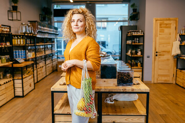 Minimalist vegan eco friendly woman choosing foods with cotton reusable net mesh bag on background of goods of zero waste shop.Shopping without plastic packaging in plastic free grocery store.