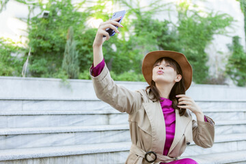 Young fashion woman dressed in autumn clothes sits on a bench and makes salfie portrait outdoors