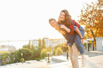 Young couple having fun in city at sunrise. Handsome man giving his girlfriend piggy back ride....
