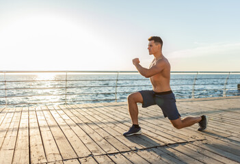 Muscular handsome man with naked torso doing lunges exercises on the beach at sunrise. Healthy lifestyle