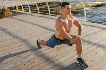 Young muscular man with naked torso doing leg stretching on the beach at sunrise