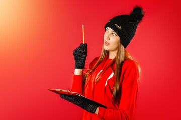 A girl in a black winter hat and gloves, a pensive look, in hands with a pencil and a list, pointing up for an advertisment