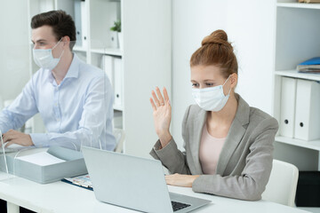 Young businesswoman in protective mask waving hand to someone on laptop display