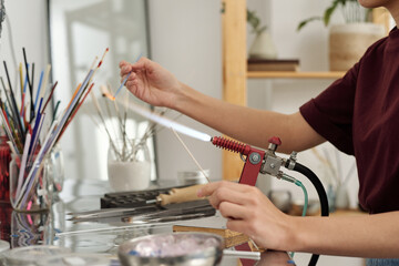 Hands of young female artisan sitting by workplace and burning glass workpiece