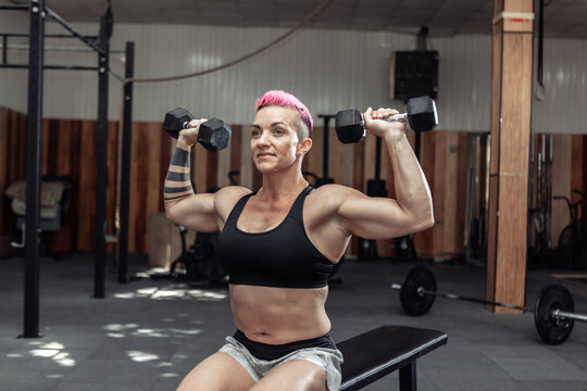 Athletic strong woman trains the muscles of the shoulder with dumbbells in her hands while sitting on a bench in a cross gym. Bodybuilding and Fitness
