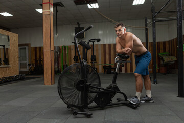 Fototapeta na wymiar Muscular tired man athlete is resting leaning his elbows on air bike in cross hall
