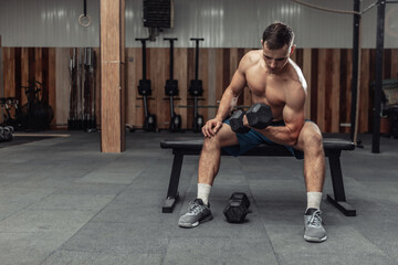 Fototapeta na wymiar Athletic man exercising with heavy dumbbell, concentrated training biceps while sitting on a bench in the gym. Bodybuilding and Fitness