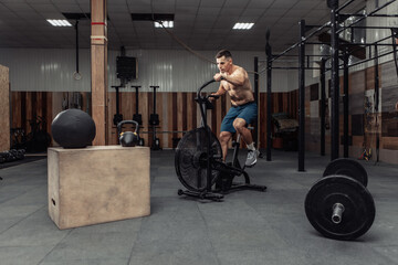 Obraz na płótnie Canvas Powerful muscular male athlete with a naked torso trains with an air bike in a modern health club. Functional, cross fit training. Cardio exercise. Healthy lifestyle concept