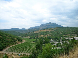 A village with houses and gardens in a picturesque valley surrounded by hills. Trees grow on the hillsides. Clouds float across the sky.