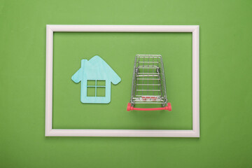 Shopping trolley with mini house in a white frame on green background. House purchase. Top view. Flat lay