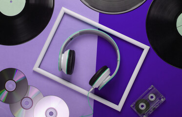 Flat lay composition from retro media, 80s. Stylish stereo headphones in white frame, vinyl records, audio cassette and cd discs on purple background. Music lover. Top view.