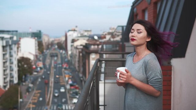 pretty young woman in stays on balcony holds cup of hot coffee or tea. Girl watching a beautiful urban view and street and relaxing alone.