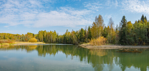 Autumn forest trees are reflected in the river water of the panoramic landscape. Blue sky with clouds.