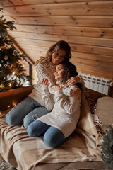 Young mother with two little daughters in pajamas decorate a New Year's tree in the cozy room with with big light window