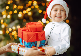 Fototapeta na wymiar Happy child in Santa red hat receives Christmas gifts. Funny little boy posing with presents in Christmas interior design. Winter holiday, New Year.