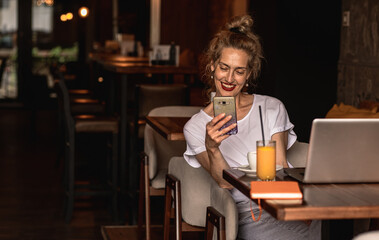 Smiling senior female blogger in casual wear sitting in coffee shop using smartphone.