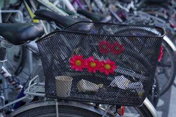 Bicycle with metal wire basket decorated with red plastic flowers. In the basket there are empty paper coffee cups. A detail of crowded bicycle parking lot at train station in Lucerne, Switzerland.