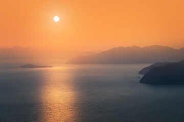 Fototapeta premium Moody sunset, the distant sun sets behind the silhouettes of blue mountains on the horizon, washed by the waters of the Mediterranean sea