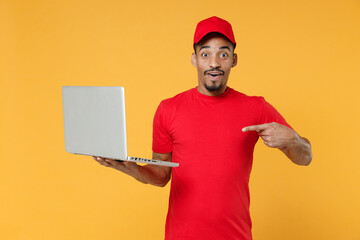 Smiling delivery employee african man in red cap blank print t-shirt uniform workwear work courier dealer service concept hold in hand using laptop pc computer isolated on yellow background studio
