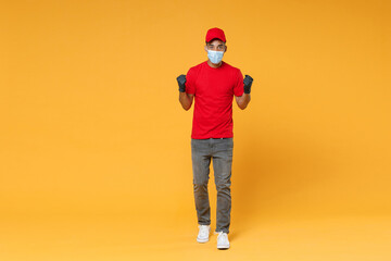 Full length delivery employee african man in red cap t-shirt face mask gloves uniform work courier dealer service on quarantine coronavirus covid-19 virus concept isolated on yellow background studio.