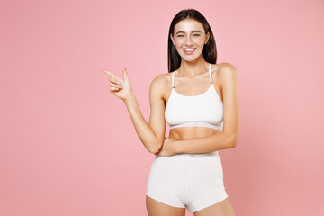 Smiling cheerful young brunette woman 20s in white underwear demonstrating beautiful body standing posing pointing index finger aside isolated on pastel pink colour wall background studio portrait.