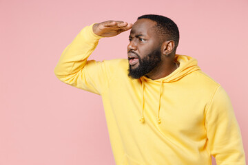 Perplexed young african american man 20s wearing basic yellow streetwear hoodie standing holding hand at forehead looking far away distance isolated on pastel pink color background studio portrait.