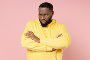 Upset offended young african american man 20s wearing casual basic yellow streetwear hoodie standing holding hands crossed looking camera isolated on pastel pink color wall background studio portrait.