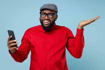 Fototapeta premium Amazed young bearded african american man 20s in casual red shirt eyeglasses cap spreading hands using mobile cell phone typing sms message isolated on pastel blue color background studio portrait.