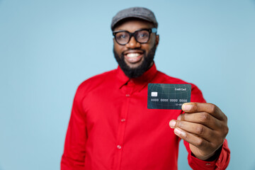 Smiling young bearded african american man 20s wearing casual red shirt eyeglasses cap standing hold credit bank card looking camera isolated on pastel blue color wall background studio portrait.