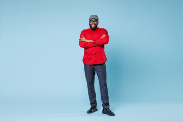 Full length of smiling handsome young bearded african american man in casual red shirt cap eyeglasses standing holding hands crossed looking camera isolated on pastel blue background studio portrait. - obrazy, fototapety, plakaty