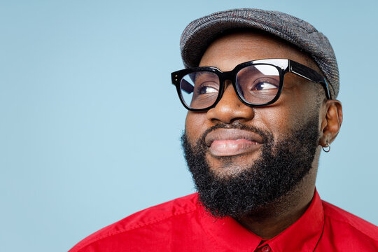 Close up of smiling pretty young bearded african american man 20s wearing casual red shirt cap eyeglasses standing and looking aside isolated on pastel blue color wall background studio portrait.