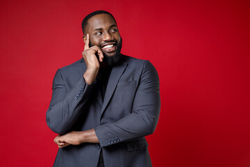 Smiling funny pensive young african american business man 20s wearing classic jacket suit standing put hand prop up on chin looking aside isolated on bright red color wall background studio portrait.