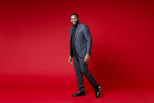 Full length side view of smiling handsome confident young african american business man 20s wearing classic jacket suit standing looking camera isolated on bright red color background studio portrait.