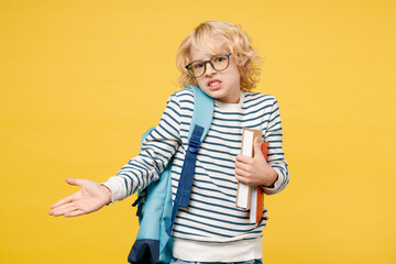 Confused little male kid teen boy 10s years old wearing striped sweatshirt eyeglasses backpack hold school books spreading hands isolated on yellow background child studio portrait. Education concept.