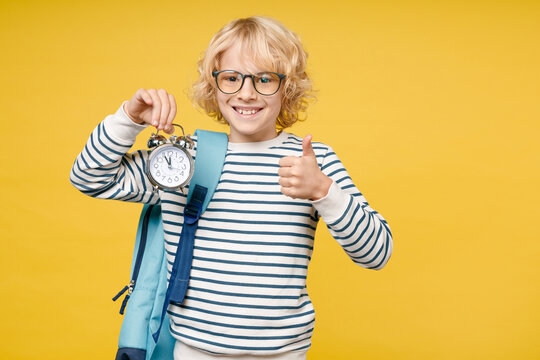 Smiling male kid teen boy 10s years old wearing striped sweatshirt eyeglasses backpack hold alarm clock showing thumb up isolated on yellow color background child studio portrait. Education concept.