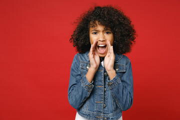 Fototapeta na wymiar Shocked little african american kid girl 12-13 years old in casual denim jacket screaming with hands gesture near mouth isolated on red color background children portrait. Childhood lifestyle concept.