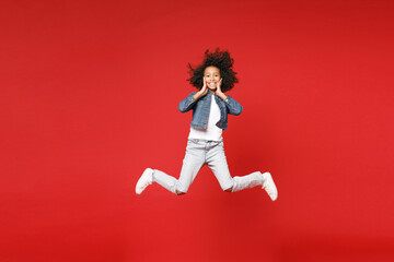 Fototapeta na wymiar Full length of smiling little african american kid girl 12-13 years old in denim jacket jumping put hands on cheeks isolated on red background children studio portrait. Childhood lifestyle concept.