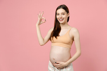 Happy young pregnant woman in basic top stroking keeping hands on belly stomach tummy with baby showing okay isolated on pastel pink background studio. Maternity family pregnancy gynecology concept