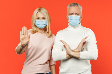 Couple two friends elderly gray-haired man blonde woman in sterile face mask to safe from coronavirus virus covid-19 showing stop gesture with palm crossed hands isolated on orange background studio.