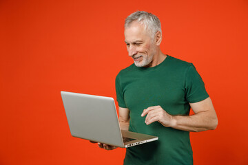 Smiling cheerful funny elderly gray-haired mustache bearded man wearing casual basic green t-shirt standing working on laptop pc computer isolated on bright orange color background studio portrait.