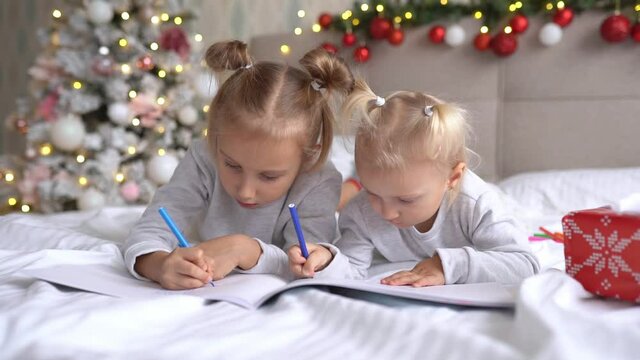 Two cute little girls sisters wearing Santa Helper hats write letter to Santa lying on bed at home in Christmas decorated room