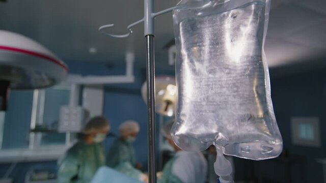 Closeup of full bag of saline for operation and team of surgeons getting ready for procedure in background