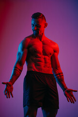 A muscular man with a beard is flexing his muscles under the blue and red light. An athletic guy is demonstrating his sporty torso. An athlete in shorts after a workout.