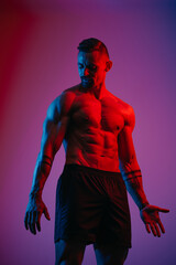 Fototapeta na wymiar A bodybuilder with a beard is flexing his muscles under the blue and red light. An athletic guy is demonstrating his sporty torso. An athlete in shorts after a workout.